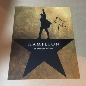 May include: A promotional poster for the musical "Hamilton" featuring a silhouette of a man on a black star. The poster has a gold and brown background with the musical's title and the words "An American Musical". Includes a signature.