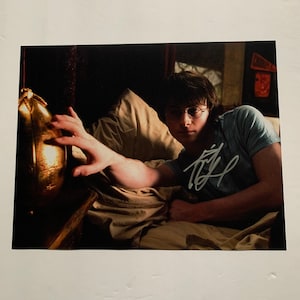 Puede incluir: Primer plano de un joven con cabello castaño y gafas acostado en la cama, alcanzando un objeto dorado con forma de huevo. La imagen está firmada "Daniel Radcliffe".