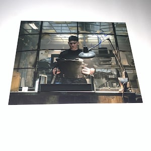 May include: A signed photograph featuring a man in a black shirt and tactical vest, holding an object. The background includes a window and a desk with a lamp. The signature is in blue ink.