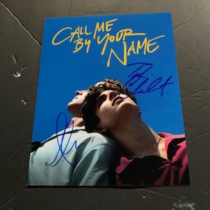 May include: A signed movie poster for the film "Call Me By Your Name" featuring two men against a blue sky background. The poster is signed by the actors Timothée Chalamet and Armie Hammer.