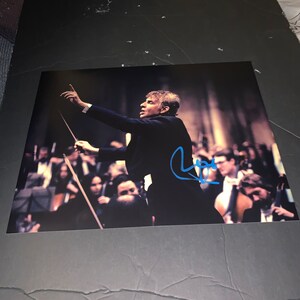 May include: A black and white photo of a conductor leading an orchestra. The conductor is wearing a black suit and is raising his baton in the air. The orchestra is seated in the background. The photo is signed by the conductor.