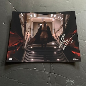 Puede incluir: Una foto en blanco y negro de un hombre con una túnica oscura bajando por una escalera. La foto está firmada "Ewan" con un pequeño signo más. La foto está etiquetada "Star Wars" y "Topps Authentic's".