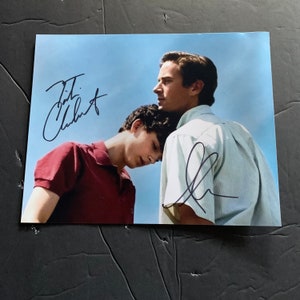 Op de afbeelding: Een gesigneerde foto van twee mannen, een met zijn arm om de ander. De man vooraan draagt een rood shirt en de man achteraan draagt een wit shirt. De handtekening "Tim Chalamet" is zichtbaar in de linkerbovenhoek van de afbeelding. De handtekening "Elio" is zichtbaar in de rechterbenedenhoek van de afbeelding.