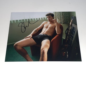 Könnte beinhalten: Ein Schwarzweißfoto einer Person, die auf einem Stuhl sitzt und schwarze Unterwäsche mit dem Schriftzug "Calvin Klein" am Bund trägt. Die Person ist oben ohne und hat ein Tattoo am linken Arm. Die Person schaut in die Kamera. Im Hintergrund befindet sich eine Tastatur.