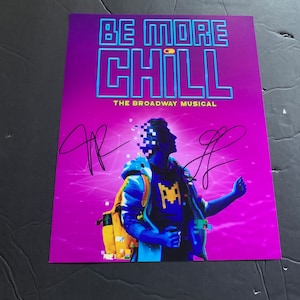 May include: A purple poster with the title "Be More Chill: The Broadway Musical" in blue and yellow letters. A pixelated image of a person wearing a blue shirt with a yellow pixelated video game character on it is in the center of the poster. The poster is signed in black ink.