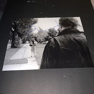 May include: Black and white photograph of a man and a woman standing on a sidewalk. The man is wearing a jacket and the woman is wearing a dress. The man is looking at the woman and the woman is looking at the camera. The photograph is signed "Clint"