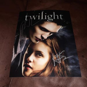 Puede incluir: Una fotografía en blanco y negro de dos personas, un hombre y una mujer, con la palabra "twilight" impresa en la parte superior. El hombre se inclina hacia la mujer, y ambos tienen sus nombres firmados con tinta plateada.