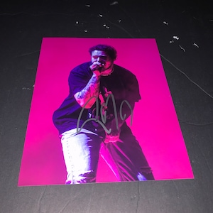 May include: A black and white photo of a person singing into a microphone. The person is wearing a black t-shirt, blue jeans, and has a black signature on the photo. The background is pink.