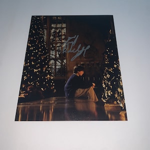 May include: A black and white photo of a young boy sitting in a large room with two decorated Christmas trees. The boy is wearing a blue sweater and khaki pants. The photo is signed "Daniel Radcliffe".