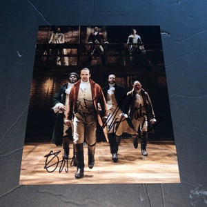 May include: A printed photograph featuring a cast of actors in period costumes, likely from a stage production. The image is signed by multiple individuals. The actors are in a theatrical setting, with a wooden stage and backdrop visible.