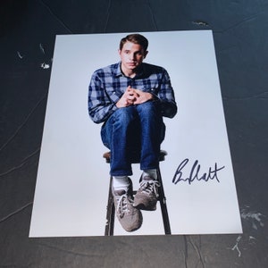 May include: A signed photograph of a man wearing a blue and white checked shirt, blue jeans, and brown shoes. He is sitting on a stool with his hands clasped in his lap. The signature is "B. J. Novak".