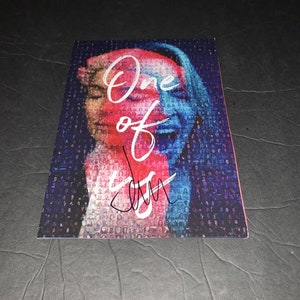 May include: A close-up of a colorful mosaic image of a person's face with the words "One of us" written in white. The image is signed with a black signature.