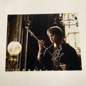 Puede incluir: Una fotografía en blanco y negro de un niño pequeño sosteniendo una varita mágica. El niño lleva una túnica oscura y gafas. La fotografía está firmada por el niño.