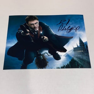 May include: A signed photograph of Daniel Radcliffe as Harry Potter flying on a broomstick in front of Hogwarts castle. The signature is in blue ink and reads "Daniel Radcliffe".