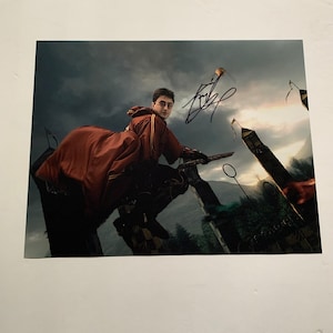 Puede incluir: Fotografía firmada que muestra a una persona montada en una escoba en una escena de Quidditch. La persona lleva un uniforme granate y dorado. El fondo muestra un cielo nublado y estructuras rotas. La fotografía está firmada con tinta negra.