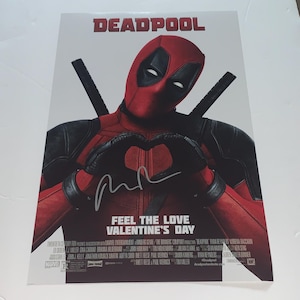 May include: A red and black Deadpool movie poster featuring Deadpool making a heart shape with his hands. The poster has the text "DEADPOOL" and "FEEL THE LOVE VALENTINE'S DAY".