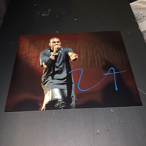 May include: A black and white photo of a man singing into a microphone on stage. The man is wearing a black shirt and pants. The photo is signed with a blue signature that says "Ye".