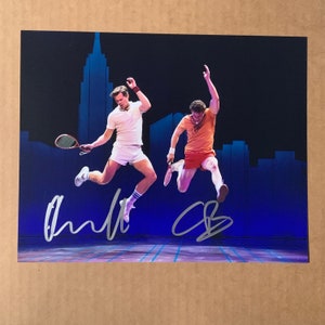Puede incluir: Una fotografía firmada de dos hombres en el aire, jugando al tenis. Los hombres llevan pantalones cortos y camisas blancas y naranjas. El fondo es un escenario azul con un telón de fondo de ciudad. Las firmas de los hombres son visibles en las esquinas inferiores izquierda y derecha de la foto.