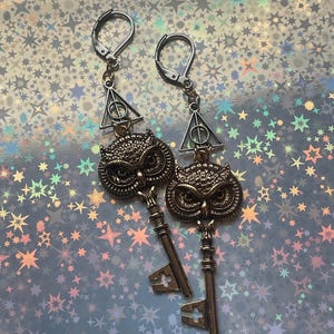 May include: A pair of gold-toned owl key earrings with a silver triangle charm featuring a symbol. The earrings have silver hooks.