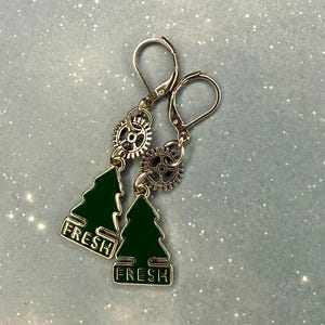 K&ouml;nnte beinhalten: Ohrringe mit gr&uuml;nen Emaille-Weihnachtsbaum-Anh&auml;ngern und dem Wort "FRESH" in Wei&szlig;. Die Ohrringe haben silberfarbene Klappb&uuml;gelverschl&uuml;sse und sind mit kleinen Zahnrad-Anh&auml;ngern verziert.