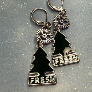 K&ouml;nnte beinhalten: Ein Paar silberne Ohrringe mit B&uuml;gelverschluss. Jeder Ohrring hat ein kleines silbernes Zahnrad, einen dunkelgr&uuml;nen Weihnachtsbaum-Anh&auml;nger und das Wort "FRESH" in wei&szlig;en Buchstaben. Die Ohrringe werden auf hellblauem Hintergrund pr&auml;sentiert.