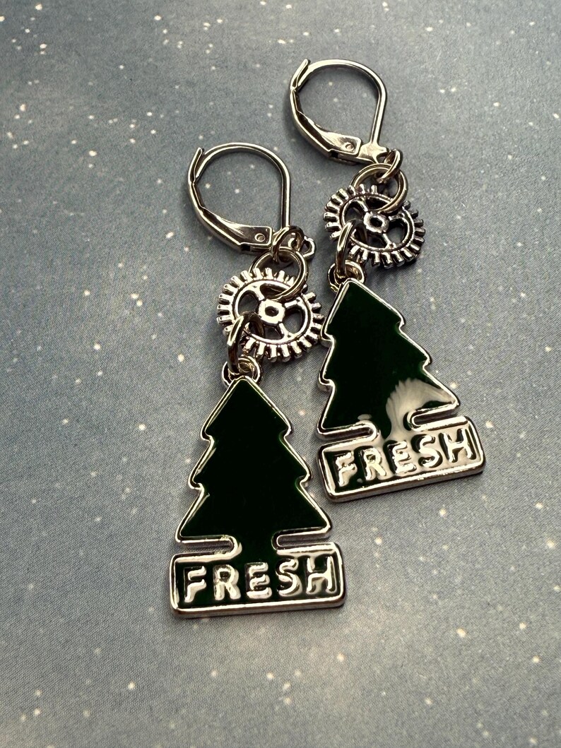 K&ouml;nnte beinhalten: Ein Paar silberne Ohrringe mit B&uuml;gelverschluss. Jeder Ohrring hat ein kleines silbernes Zahnrad, einen dunkelgr&uuml;nen Weihnachtsbaum-Anh&auml;nger und das Wort "FRESH" in wei&szlig;en Buchstaben. Die Ohrringe werden auf hellblauem Hintergrund pr&auml;sentiert.