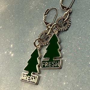 K&ouml;nnte beinhalten: Silberfarbene Ohrringe mit Klappb&uuml;gelverschl&uuml;ssen. Jeder Ohrring hat einen gr&uuml;nen Emaille-Weihnachtsbaum-Anh&auml;nger mit dem Wort "FRESH" in Wei&szlig;. Kleine zahnradf&ouml;rmige Akzente sind ebenfalls enthalten.