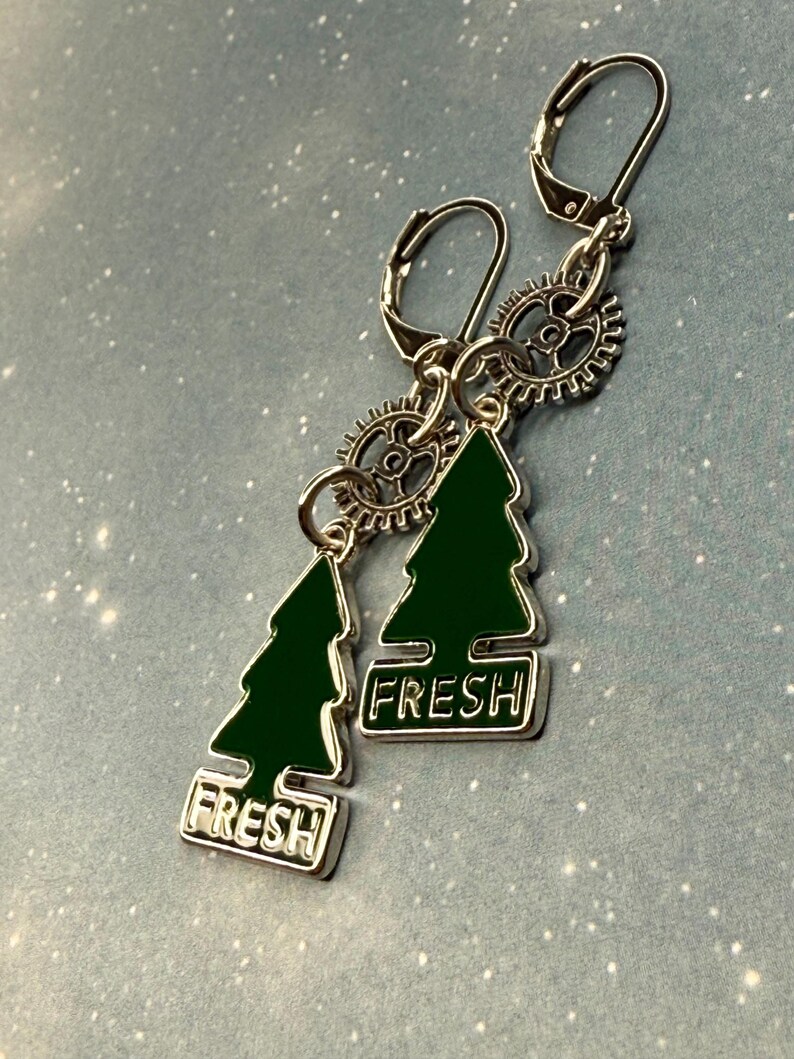 K&ouml;nnte beinhalten: Silberfarbene Ohrringe mit Klappb&uuml;gelverschl&uuml;ssen. Jeder Ohrring hat einen gr&uuml;nen Emaille-Weihnachtsbaum-Anh&auml;nger mit dem Wort "FRESH" in Wei&szlig;. Kleine zahnradf&ouml;rmige Akzente sind ebenfalls enthalten.