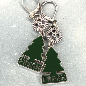 K&ouml;nnte beinhalten: Ein Paar silberfarbene Ohrringe mit B&uuml;gelverschluss. Jeder Ohrring hat einen kleinen Zahnrad-Anh&auml;nger und einen gr&uuml;nen Emaille-Weihnachtsbaum-Anh&auml;nger mit dem Wort "FRESH" in wei&szlig;er Schrift. Die Ohrringe werden auf einer hellblauen Oberfl&auml;che pr&auml;sentiert.