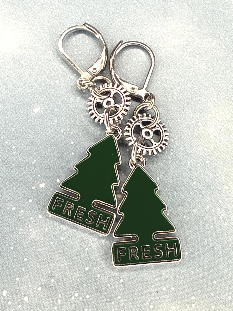 K&ouml;nnte beinhalten: Ein Paar silberfarbene Ohrringe mit B&uuml;gelverschluss. Jeder Ohrring hat einen kleinen Zahnrad-Anh&auml;nger und einen gr&uuml;nen Emaille-Weihnachtsbaum-Anh&auml;nger mit dem Wort "FRESH" in wei&szlig;er Schrift. Die Ohrringe werden auf einer hellblauen Oberfl&auml;che pr&auml;sentiert.