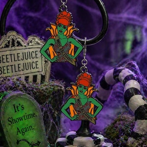 May include: A pair of dangling earrings featuring a green-skinned woman with red hair and a sash that reads "Miss Argentina." The earrings are set against a spooky, Halloween-themed backdrop with a tombstone that reads "It's Showtime. Again."