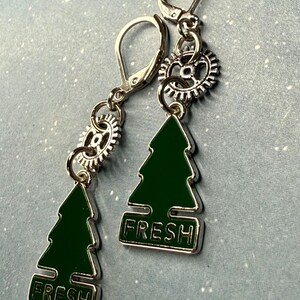 K&ouml;nnte beinhalten: Silberfarbene Ohrringe mit einem gr&uuml;nen Weihnachtsbaum-Anh&auml;nger und dem Wort "FRESH". Jeder Ohrring hat einen kleinen zahnradf&ouml;rmigen Anh&auml;nger und einen B&uuml;gelverschluss.