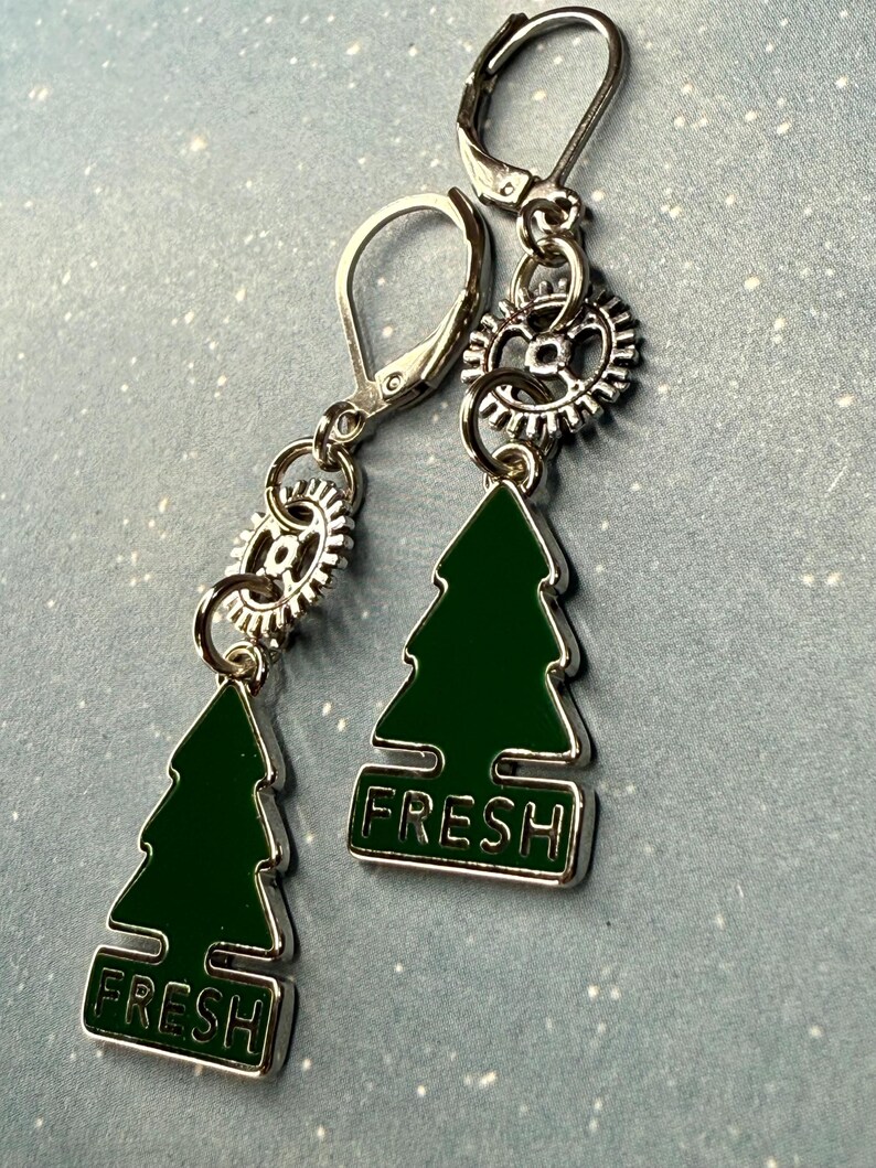 K&ouml;nnte beinhalten: Silberfarbene Ohrringe mit einem gr&uuml;nen Weihnachtsbaum-Anh&auml;nger und dem Wort "FRESH". Jeder Ohrring hat einen kleinen zahnradf&ouml;rmigen Anh&auml;nger und einen B&uuml;gelverschluss.