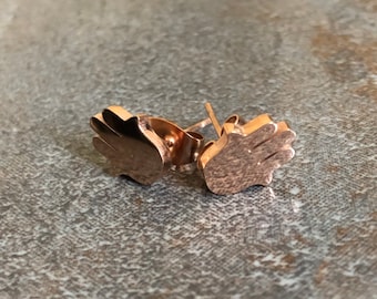 May include: A pair of rose gold stud earrings. Each earring is shaped like a hand with the fingers slightly spread. The earrings are displayed on a textured, grey surface. The earrings are small and delicate.