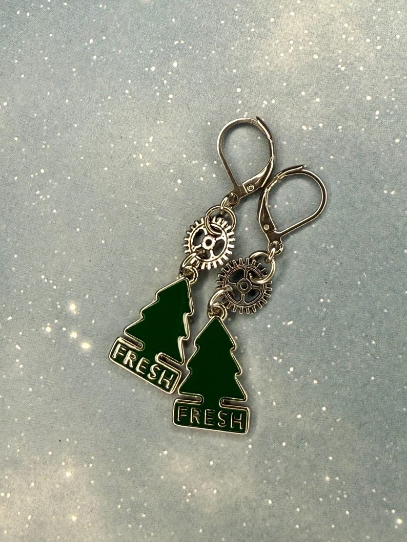 K&ouml;nnte beinhalten: Silberfarbene Ohrringe mit Steampunk-&Auml;sthetik. Jeder Ohrring hat einen kleinen, dunkelgr&uuml;nen Weihnachtsbaum-Anh&auml;nger mit dem Wort "FRESH" in wei&szlig;er Schrift. Der Baum-Anh&auml;nger h&auml;ngt unter einem silbernen Zahnrad.
