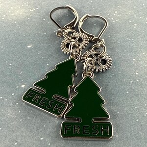 K&ouml;nnte beinhalten: Silberne Ohrringe mit B&uuml;gelverschluss. Jedes Ohrring-Paar hat einen gr&uuml;nen Emaille-Weihnachtsbaum-Anh&auml;nger mit dem Wort "FRESH". Die Ohrringe haben auch kleine silberne Zahnrad-Anh&auml;nger.