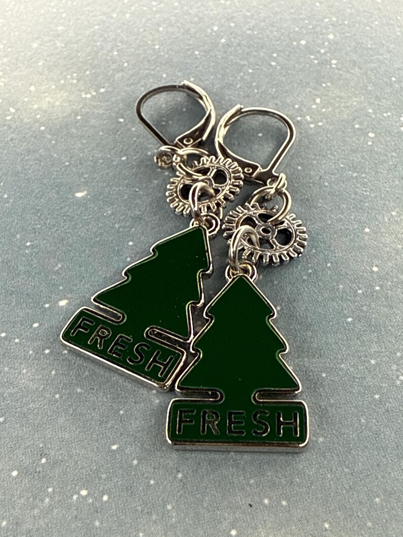 K&ouml;nnte beinhalten: Silberne Ohrringe mit B&uuml;gelverschluss. Jedes Ohrring-Paar hat einen gr&uuml;nen Emaille-Weihnachtsbaum-Anh&auml;nger mit dem Wort "FRESH". Die Ohrringe haben auch kleine silberne Zahnrad-Anh&auml;nger.