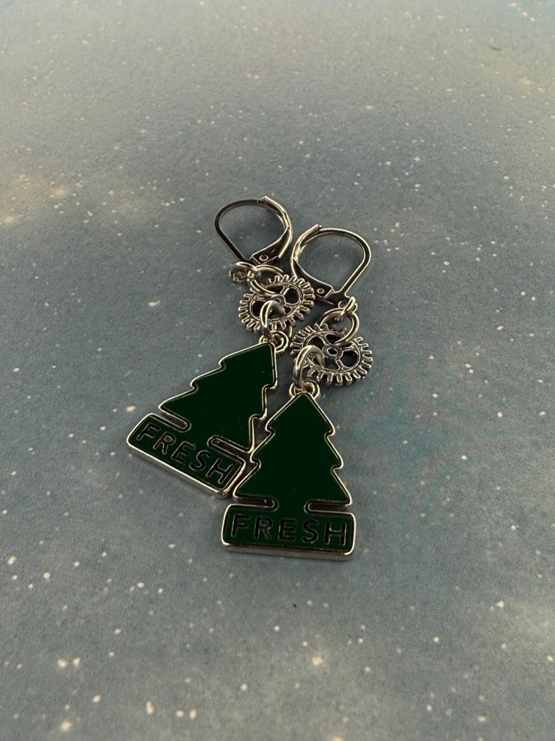 K&ouml;nnte beinhalten: Silberfarbene Ohrringe mit einem gr&uuml;nen Weihnachtsbaum-Design und dem Wort "FRESH". Die Ohrringe haben einen Klappverschluss und sind mit kleinen zahnradf&ouml;rmigen Anh&auml;ngern versehen.