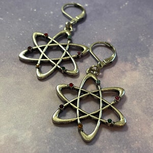 Może przedstawiać: Para srebrnych kolczyków w kształcie atomu z kolorowymi kamieniami szlachetnymi. Kolczyki mają kształt atomu z srebrną pętelką do zawieszenia.