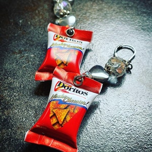 May include: A pair of earrings featuring miniature Doritos Nacho Cheese bags with silver charms and crystal beads. The bags are red with white text and a picture of nacho cheese flavored chips.