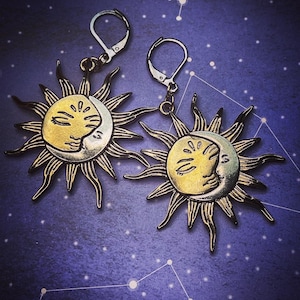 Puede incluir: Un par de pendientes de oro con un diseño de sol y luna. Los pendientes están hechos de metal y tienen un aspecto vintage.