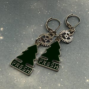 K&ouml;nnte beinhalten: Ohrringe zum H&auml;ngen mit einem dunkelgr&uuml;nen Weihnachtsbaum-Design und dem Wort "FRESH" in Wei&szlig;. Die Ohrringe haben silberfarbene Klappb&uuml;gelverschl&uuml;sse und sind mit einem zahnradf&ouml;rmigen Anh&auml;nger verbunden.