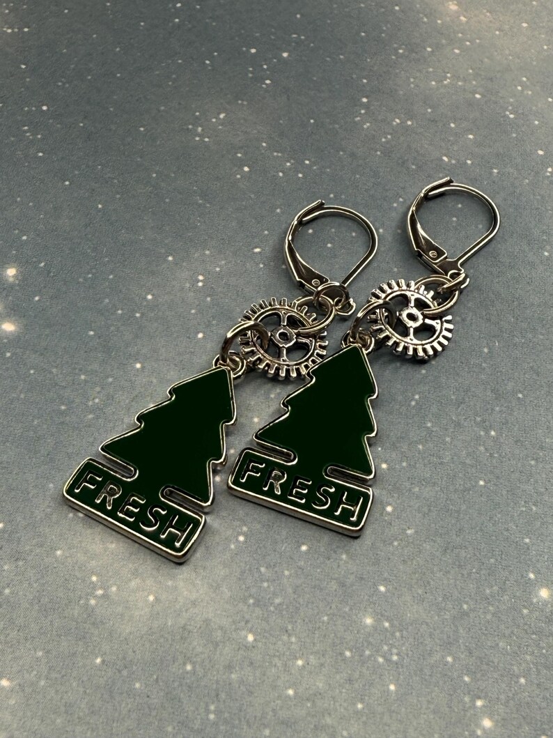 K&ouml;nnte beinhalten: Ohrringe zum H&auml;ngen mit einem dunkelgr&uuml;nen Weihnachtsbaum-Design und dem Wort "FRESH" in Wei&szlig;. Die Ohrringe haben silberfarbene Klappb&uuml;gelverschl&uuml;sse und sind mit einem zahnradf&ouml;rmigen Anh&auml;nger verbunden.
