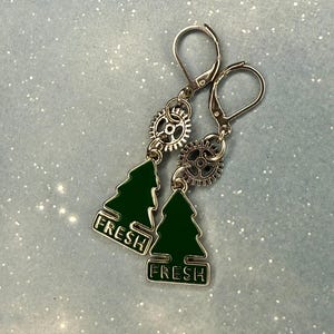 K&ouml;nnte beinhalten: Ein Paar silberfarbene Ohrringe mit Klappb&uuml;gelverschl&uuml;ssen. Jeder Ohrring hat einen gr&uuml;nen Emaille-Weihnachtsbaum-Anh&auml;nger mit dem Wort "FRESH" in Wei&szlig; und ein kleines zahnradf&ouml;rmiges Detail. Die Ohrringe sind vor einem hellblauen Hintergrund platziert.