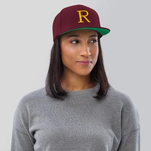 May include: A maroon baseball cap with a green underside and a gold embroidered letter "R" on the front.