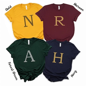 May include: Four t-shirts in different colors with a letter embroidered on each. The shirts are gold, maroon, forest green, and navy. The letters are N, R, A, and H.