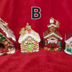 K&ouml;nnte beinhalten: Vier gl&auml;serne Weihnachtsbaumschmuckst&uuml;cke in Form von H&auml;usern. Die Schmuckst&uuml;cke sind mit Glitzer, Zuckerstangen und anderen festlichen Details verziert. Die H&auml;user umfassen ein Lebkuchenhaus, ein Schulhaus, eine Kirche und ein Hundehaus.