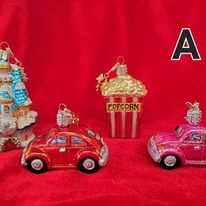 K&ouml;nnte beinhalten: Vier gl&auml;serne Weihnachtsbaumschmuckst&uuml;cke, darunter ein blaues und wei&szlig;es Schloss, ein roter Volkswagen K&auml;fer, ein rosa Volkswagen K&auml;fer und ein Popcorn-Eimer mit roten und wei&szlig;en Streifen. Der Schmuck befindet sich auf einem roten Samt-Hintergrund.