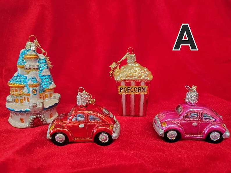 K&ouml;nnte beinhalten: Vier gl&auml;serne Weihnachtsbaumschmuckst&uuml;cke, darunter ein blaues und wei&szlig;es Schloss, ein roter Volkswagen K&auml;fer, ein rosa Volkswagen K&auml;fer und ein Popcorn-Eimer mit roten und wei&szlig;en Streifen. Der Schmuck befindet sich auf einem roten Samt-Hintergrund.