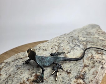 May include: A bronze lizard sculpture with a dark green and gold finish. The lizard is posed on a speckled stone. The sculpture has detailed legs and a long tail. The lizard is a decorative item.